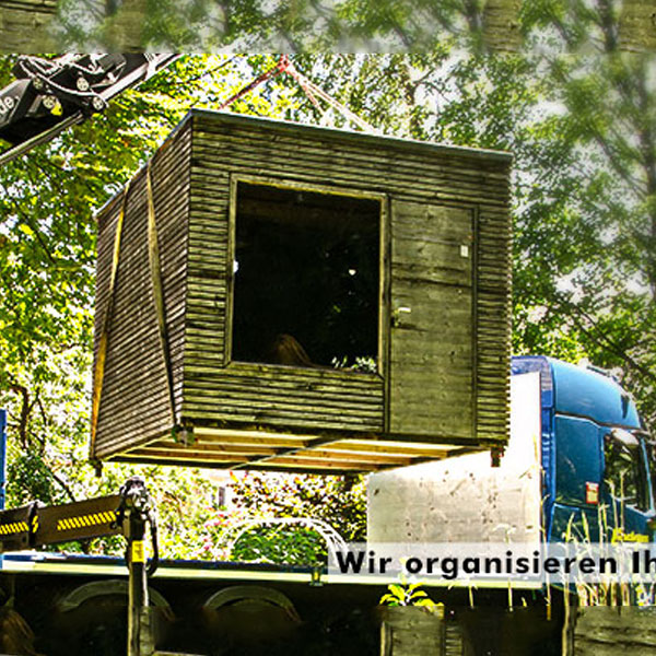 Ein Kran hebt ein hölzernes Gartenhaus auf einen Endras-Tieflader